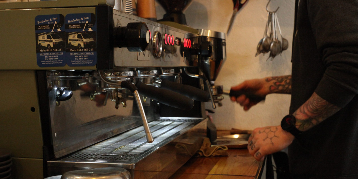 A tattooed man brews coffee in a busy shop, ensuring quality and consistency for customers' daily cups.