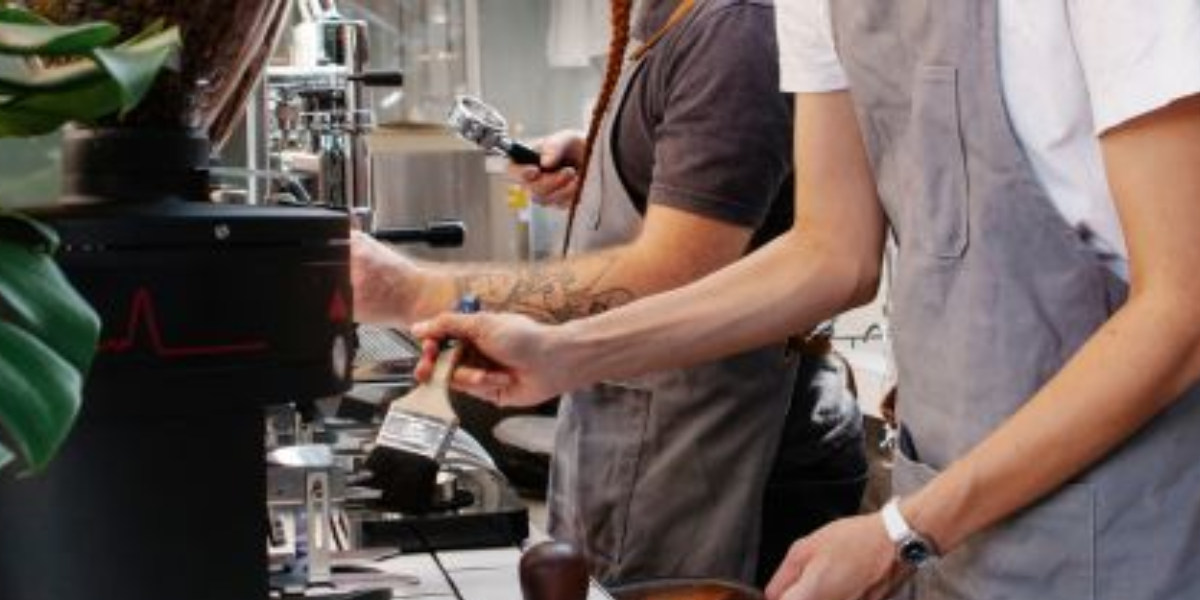Two people working on laptops at a coffee shop, surrounded by coffee equipment and a cozy atmosphere.